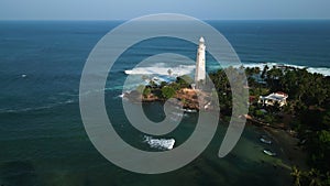 Drone flies over Dondra Head Lighthouse surrounded by trees, ocean waves crashing on rocks, clear blue sea merging with