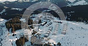 Drone flies up from beautiful idyllic winter resort village among sunny morning landscape, snow covered mountains.