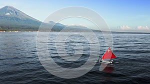 Drone flyby about scarlet red sail white canoe boat floating on ocean water near sea shore with huge volcano. Idyllic