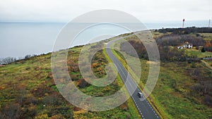 Drone flying above black car moving along clear road near beautiful lighthouse sea bay and autumn field on cloudy day.