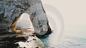 Drone flying right along epic natural rock arch at famous white alabaster seaside cliffs at Etretat Normandy on sunset.