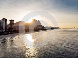 Drone flying towards Barra da Tijuca beach while filming waves crashing on beach during sunrise, with the buildings and