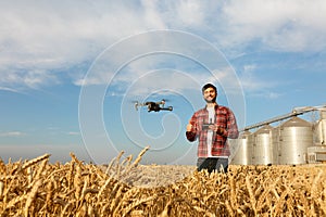 Drone hovers in front of farmer with remote controller in hands near grain elevator. Quadcopter flies near pilot