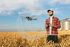 Drone hovers in front of farmer with remote controller in hands near grain elevator. Quadcopter flies near pilot