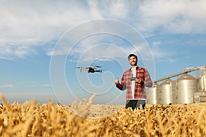 Drone hovers in front of farmer with remote controller in hands near grain elevator. Quadcopter flies near pilot