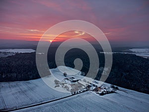 Drone panorama of amazing red sunrise behind clouds over bavarian forest and isolated farm area with snow in winter