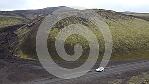 Drone shot of a car going by a green moss-covered volcanic mountain and black lava field, Iceland