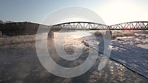 Drone Shot, Flies Backward Over the Frozen River at Sunrise, Car Driving on the Red Steel Bridge, Aerial 4k Footage