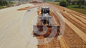 Drone shot, front view of a giant yellow bulldozer during a construction of a new highway, moving loose sand.