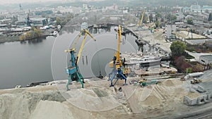 Drone shot of industrial city in fog and smog with construction and cargo cranes unloading iron barge and scow near pile of sand