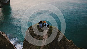 Drone tilts down over happy romantic couple watching sunset ocean view sitting on top of famous Normandy coast cliff.