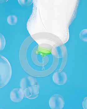 A drop of dishwashing gel flows out of a bottle on a blue background with flying soap bubbles.