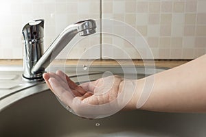 A drop of water drips from faucet in human palm above round metal kitchen sink