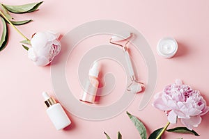 Dropper bottles with cosmetic oil, serum, facial roller, cream jar and peony flowers on pink background. Top view, flat photo