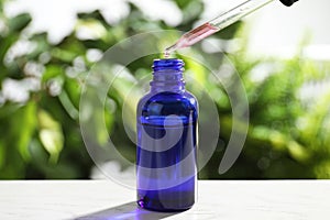 Dropper with essential oil over bottle on white wooden table against blurred