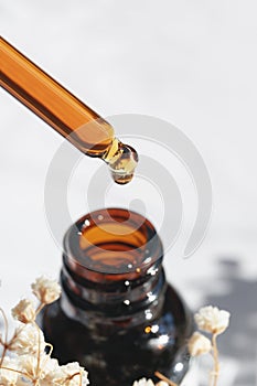 Dropper pipette with serum or oil and amber bottle. Skincare products, natural cosmetic and flowers on white background.