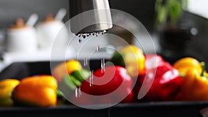 Drops of water drip from the faucet into the sink against the background of red and yellow peppers in slow motion. The