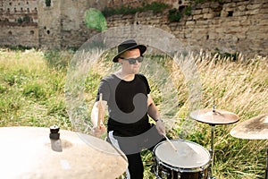 Drummer in black hat rehearses in the open air. He plays drums in the grass near the old castle