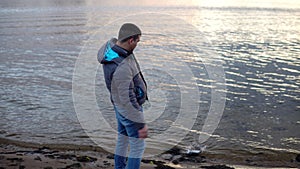 A drunk man in a jacket walks along the river bank and throws a can into the river. Environmental pollution