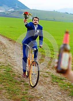 Drunken groom on a bike holding a wedding bouquet is running after a bride with a beer bottle.