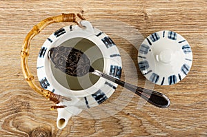 Dry black tea in spoon above opened teapot, cap on wooden table. Top view