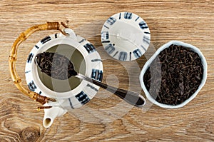 Dry black tea in spoon above striped teapot, cap, tea in bowl on wooden table. Top view