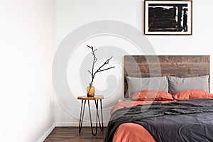 Dry branch in glass vase on wooden nightstand table nest to king size bed with orange and grey bedding