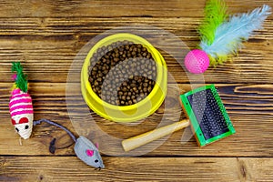 Dry cat food in bowl, cat toys and pet slicker brush on wooden background. Top view. Pet care concept