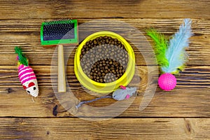 Dry cat food in bowl, cat toys and pet slicker brush on wooden background. Top view. Pet care concept