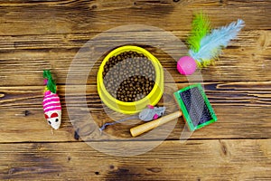 Dry cat food in bowl, cat toys and pet slicker brush on wooden background. Top view. Pet care concept