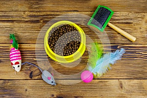 Dry cat food in bowl, cat toys and pet slicker brush on wooden background. Top view. Pet care concept