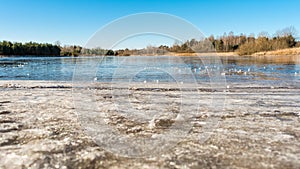 Dry grass is covered with hoarfrost. Lake shore with thawed water and ice patches on a frosty spring day. Sunny day with clear sky