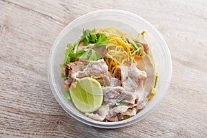 Dry noodles pork with red roasted pork and vegetable in plastic bowl on wooden table, top view Thai and China Asian food yellow