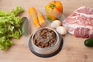 Dry pet dog food with natural ingredients. Raw meat, vegetables, eggs and salad near bowl with dry pet feed on wooden background.