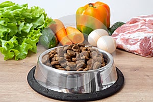 Dry pet dog food with natural ingredients. Raw meat, vegetables, eggs and salad near bowl with dry pet feed on wooden background.