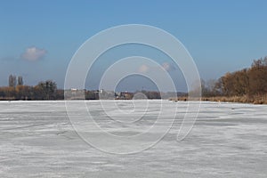 Dry reeds and trees are on the shore of a lake covered with ice