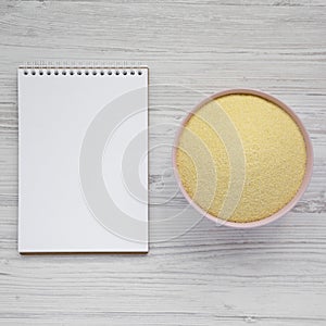 Dry semolina durum flour in a pink bowl, blank notepad over white wooden surface, top view. Overhead, from above, flat lay. Space