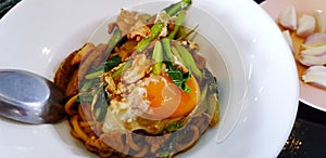 Dry Thai noodle with seafood, kale and yolk on top on white plate or dish with blurred garlic on pink dish.