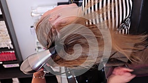 Drying blond hair with hair dryer and round brush at hairdresser. Vertical 4K.