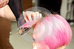 Drying short pink hair of young caucasian woman with a black hairdryer and black round brush by hands of a male