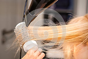 Drying straight blond hair with black hairdryer and white round brush in hairdresser salon, close up.