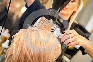 Drying straight blond hair with black hairdryer and white round brush in hairdresser salon, close up.