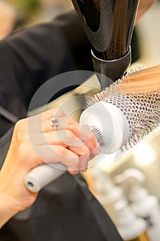 Drying straight blond hair with black hairdryer and white round brush in hairdresser salon, close up.