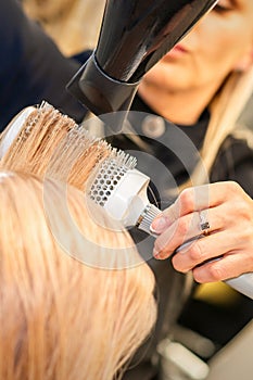 Drying straight blond hair with black hairdryer and white round brush in hairdresser salon, close up.