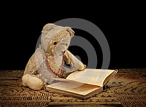 Old Teddy bear reading an old book, black background.
