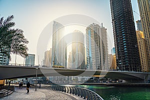 Dubai Marina promenade with walking path, canal with boats and high buildings in sunlight, United Arab Emirates, Middle