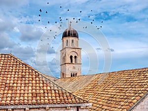 A Dubrovnik Old Town bell tower and tiled roof tops, as birds fly through the sky.