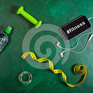 Dumbbell, water bottle, smartphone, headphones and measuring tape on green background. Hashtag fitness on the smartphone