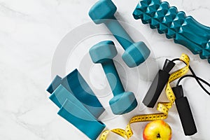 Dumbbells, elastic band, jump rope, roller, apple, measure tape on marble background. Female workout training equipment top view.