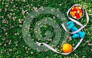 Dumbbells, measuring tape and fruit salad on green grass. Top view, copy space